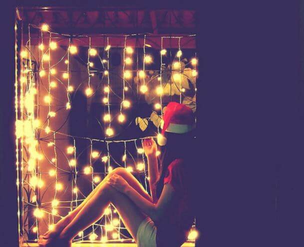 Dziewczyna w czapce Mikołaja siedzi przy lampkach świątecznych.